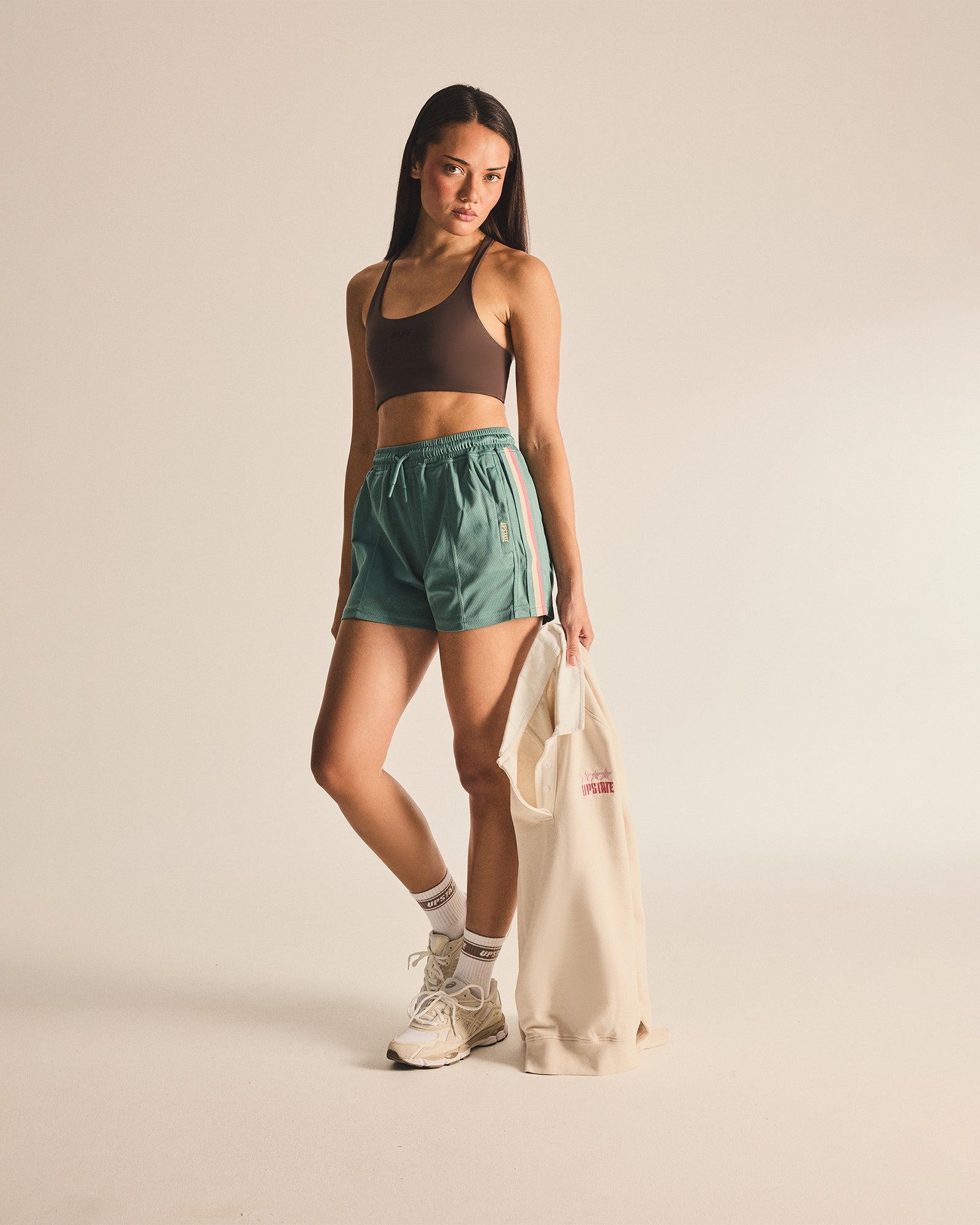 Woman wearing a brown sports bra and green shorts, holding a beige tote bag on a beige background