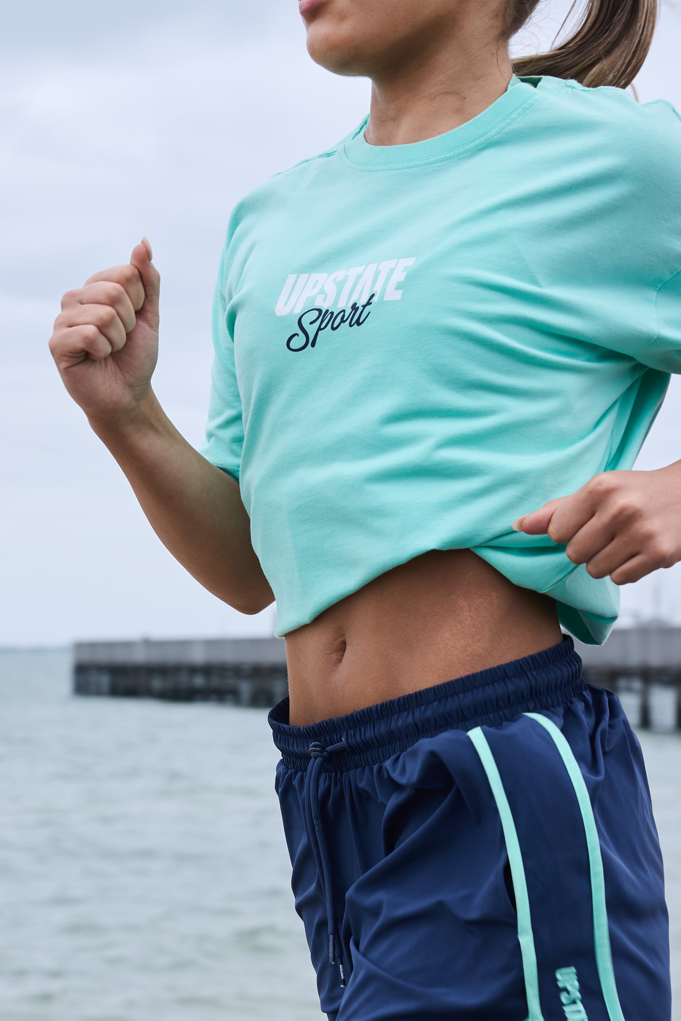 Person wearing a light blue athletic shirt and navy shorts with a white stripe, standing by water.